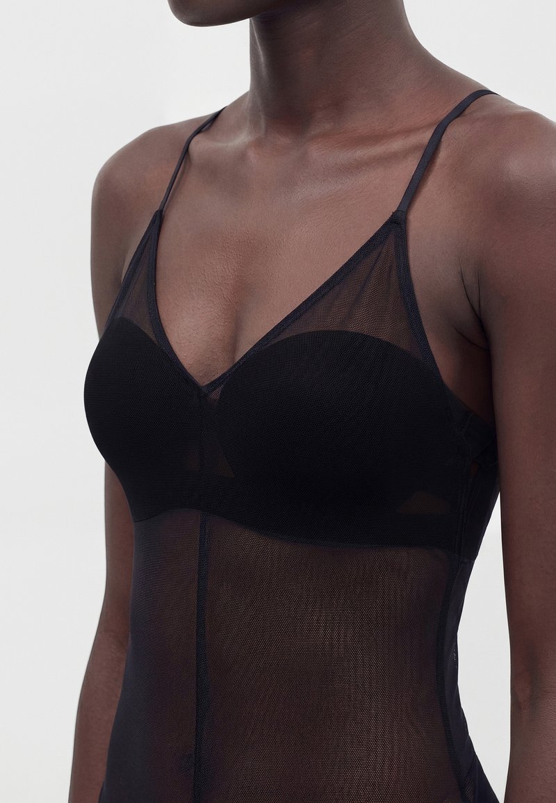Femme portant un caraco en mesh noir transparent avec des fines bretelles, montrant des bonnets de soutien-gorge intégrés et un tissu lisse ajusté sur le torse.