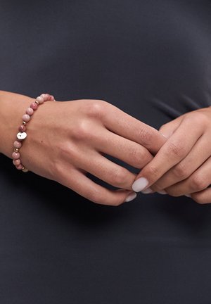 Hände mit hellrosa Nagellack, die ein rosa Perlenarmband mit goldenen Akzenten und einem kleinen goldenen Anhänger auf einem dunklen Stoffhintergrund tragen.