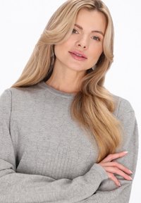 Lichtgrijze gebreide trui met een structuurpatroon, geribde halslijn en manchetten. Model heeft lang blond haar en poseert nonchalant met gekruiste armen.