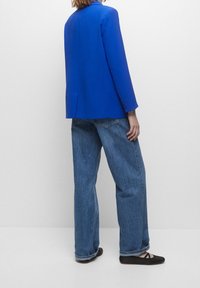 Blazer bleu cintré, jean en denim ample et chaussures plates noires. Présente un tissu lisse et une silhouette structurée avec des boutons décoratifs sur les manches.