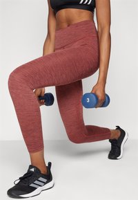 Leggings de sport rouge marbré avec une taille haute, associés à un soutien-gorge de sport noir et à des chaussures de sport noires, tenant des haltères bleus.