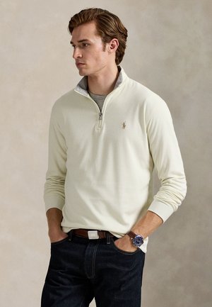 Un hombre luce un jersey color crema con cremallera corta, vaqueros oscuros y un reloj de pulsera. Está de pie con las manos en los bolsillos ante un fondo neutro.