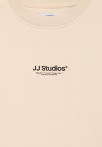 Béžová bavlněná mikina s kulatým výstřihem, na přední straně s černým logem a textem "JJ Studios". Hladká textura.