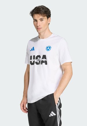 Bijela majica s kratkim rukavima s crnim natpisom "USA", plavim Adidas logom i plavim akcentom, u kombinaciji s crnim hlačama s bijelim prugama.
