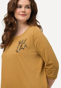 Camisa de color mostaza con mangas tres cuartos, que presenta bordados florales en blanco y negro en la parte izquierda del pecho. Textura suave.