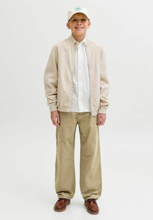 Dječak u bež jakni, bijeloj košulji, kaki hlačama, smeđim cipelama i bijelom kapom, stoji na jednostavnoj svijetloj pozadini i smiješi se.