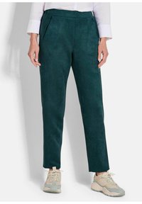 Donkergroene suède broek met een getailleerd ontwerp, voorzien van plooien aan de voorkant en zijzakken, gecombineerd met sneakers in neutrale tinten.