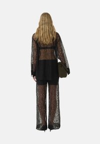 Completo in pizzo nero composto da un top a maniche lunghe con schiena scoperta e pantaloni a gamba larga. Il tessuto presenta un motivo floreale. Il modello tiene un accessorio verde.