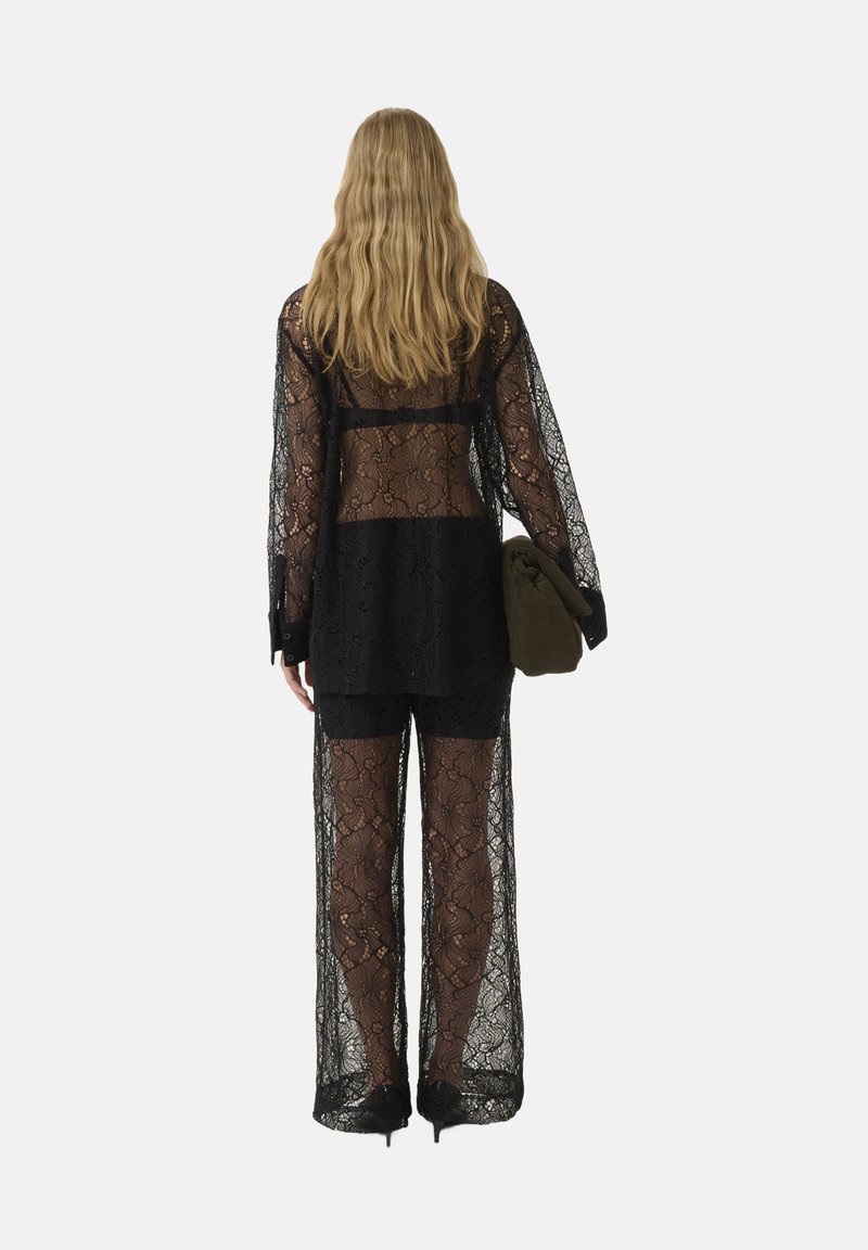 Schwarzes Spitzenoutfit mit einem langärmeligen, offenrückigen Oberteil und weit geschnittenen Hosen. Der Stoff hat ein florales Muster. Das Modell hält ein grünes Accessoire.
