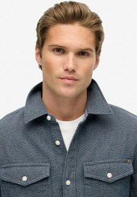 Szara, teksturowana kurtka koszulowa z wydłużonym kołnierzem, białymi guzikami i dwiema kieszeniami na piersi. Nosi się ją na białej koszuli.