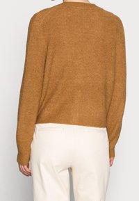 Person, der einen braunen Strickpullover und cremefarbene Hosen trägt, von hinten vor einem schlichten weißen Hintergrund gezeigt.