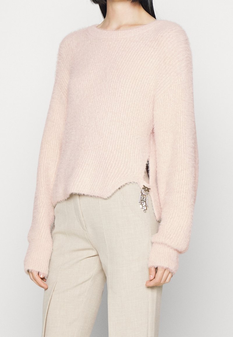 Kobieta w jasnoróżowym, puszystym swetrze z bocznym rozcięciem i detalem w postaci przywieszki, w zestawie z beżowymi spodniami o wysokim stanie.