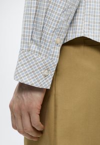 Αριστερό χέρι δίπλα σε μπεζ παντελόνια, φορώντας ένα ανοιχτό μπλε και άσπρο καρό πουκάμισο με μακρύ μανίκι και μανσέτα με δύο λευκά κουμπιά.