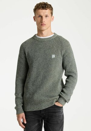 Mann mit lockigem Haar, der einen grauen Strickpullover und schwarze Jeans trägt, steht mit einer Hand in der Tasche vor einem einfarbigen weißen Hintergrund.
