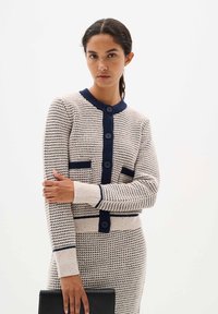 Knit cardigan in cream with navy accents, featuring a round neck, button front, and pocket details. Fitted design with textured pattern.
