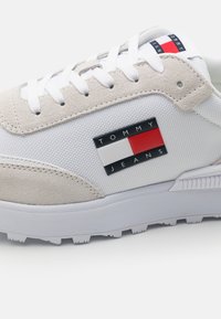 Vit sneaker med beige mocka detaljer, texturerade nät sidor och en framträdande svart, röd och vit logotyp patch. Inkluderar vita snören och gummisula.