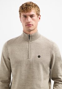 Beige Kapuzenpullover mit Reißverschluss und hohem Kragen, mit einem strukturierten Strickmuster und einem kleinen schwarzen Hexagon-Logo auf der linken Brust.
