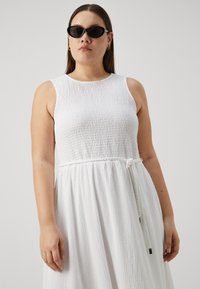 Abito bianco senza maniche con texture plissettata, corpetto aderente e dettaglio in corda intrecciata in vita. Modella che indossa occhiali da sole neri.