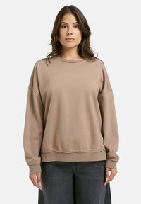 Frau mit bräunlichem, mittellangem Haar, die einen lockeren beigen Sweatshirt und dunkle graue Hosen trägt, steht vor einem schlichten hellen Hintergrund.