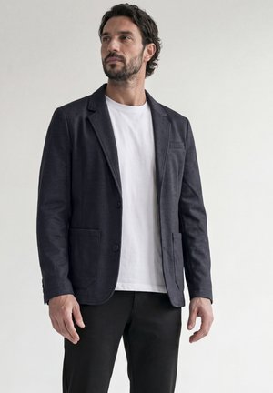 Homme debout portant un blazer sombre sur un t-shirt blanc et un pantalon noir, sur un fond clair uni.