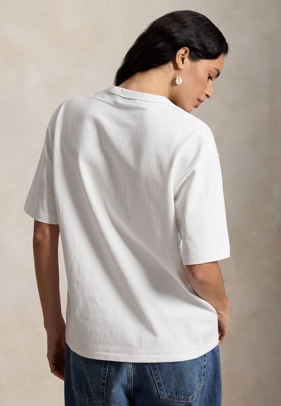 Sötét hajú nő fehér rövid ujjú pólóban és kék farmerral, háttal a néző felé, fejét oldalra fordítva.