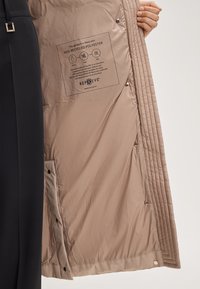 Beżowa kurtka rozpięta, ukazująca wewnętrzną metkę informującą, że wypełnienie stanowi 100% izolacja z poliestru pochodzącego z recyklingu, dla zrównoważonego rozwoju.