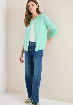 Młoda kobieta ubrana w miętowy sweter na guziki, białą koszulę, niebieskie spodnie dzwony i białe trampki z zielonymi detalami, stojąca wewnątrz pomieszczenia.