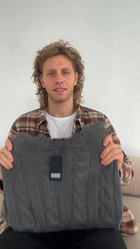 Grau gestrickter Pullover mit Kabelmustern, weicher Textur, rundem Ausschnitt und angehängtem Etikett. Modell hält ihn vor einem neutralen Hintergrund.