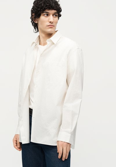 Νέος άνδρας με σγουρά σκούρα μαλλιά που φοράει ένα ανοιχτό λευκό ριγέ πουκάμισο πάνω από ένα λευκό t-shirt και μπλε τζιν, κοιτάζει αριστερά.