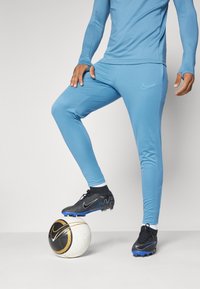 Parte superior atlética de manga larga azul clara y pantalones ajustados; botas de fútbol negras con acentos azules; de pie sobre un balón de fútbol blanco y dorado.