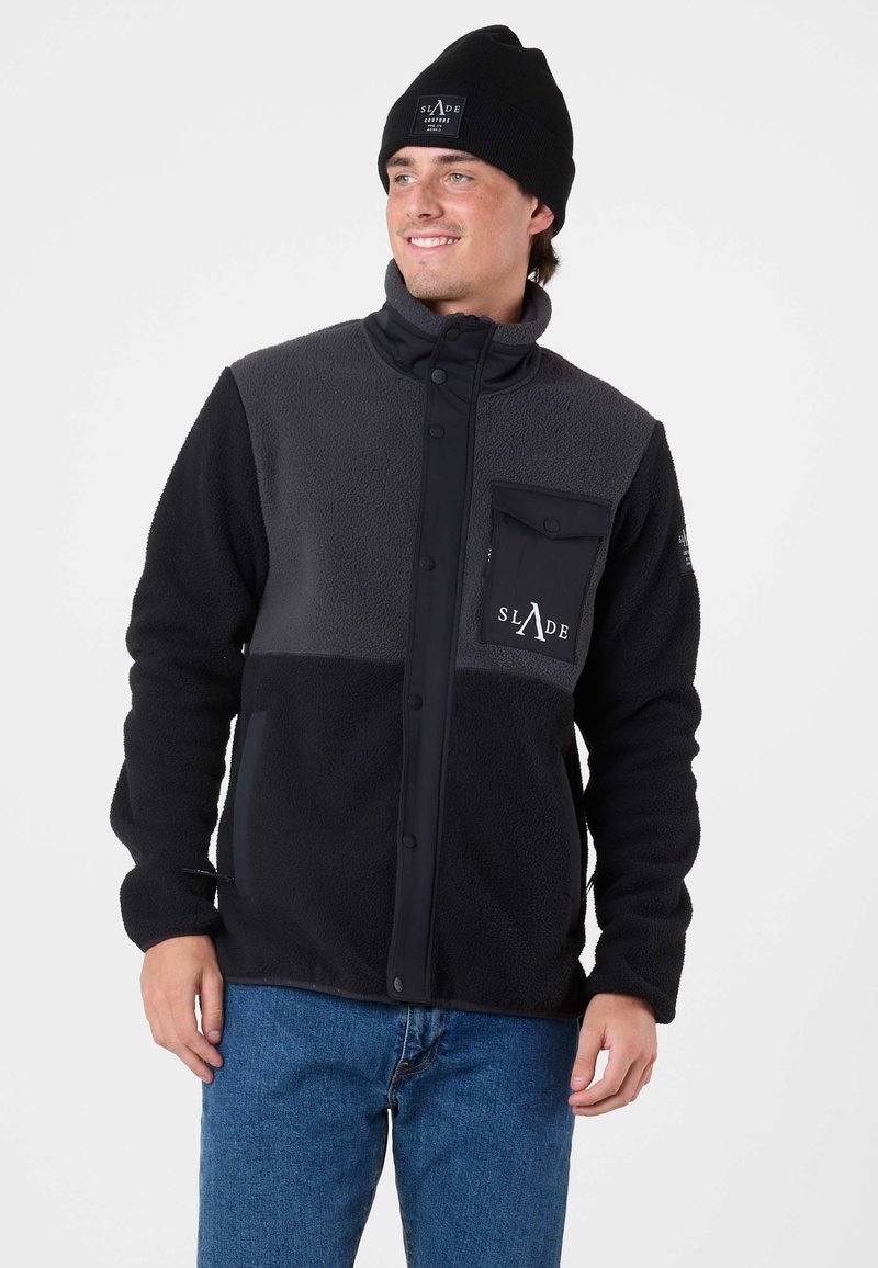 Giacca in fleece nera e grigia con chiusura a bottoni, una tasca sul petto e tessuto testurizzato. Indossata con un berretto nero.