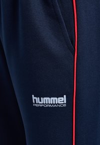 Zbliżenie na granatowe spodnie sportowe z czerwonym paskiem i logo "hummel PERFORMANCE" w pobliżu kieszeni.