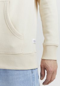 Felpa beige con tasca frontale, polsini a costine e un piccolo tag del brand sul lato inferiore; texture liscia e vestibilità comoda.