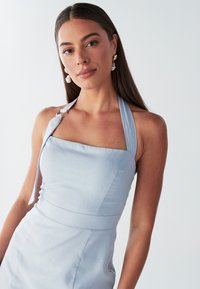 Lichtblauwe halterjurk, vierkante halslijn, getailleerde top, subtiele textuur, met een gouden accent op de schouders. Pareloorbellen maken de look compleet.
