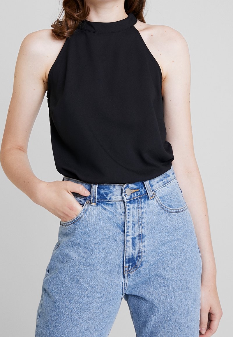 Haut noir sans manches avec un col montant et un tissu lisse, associé à un jean taille haute délavé clair avec des poches classiques et une fermeture à bouton.