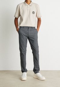 Beige polo shirt with a circular logo, paired with dark textured trousers and white sneakers. Casual outfit with a simple design.