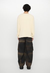 Pull à rayures de couleur crème avec des manches longues. Pantalons cargo noirs avec un motif texturé en dégradé et des poches latérales. Chaussures noires épaisses.