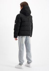 Zwarte gewatteerde jas met een capuchon, voorzien van dikke isolatie, zijzakken en een gladde textuur, gecombineerd met lichte distressed jeans en witte sneakers.