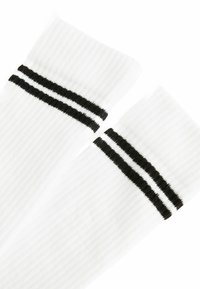 Chaussettes blanches montantes avec une texture ribbed, ornées de deux rayures noires en haut. Le design met en avant la longueur et un style sportif.