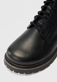 Botín de cuero negro con superficie texturizada, puntera redonda, cordones negros y suela de goma gruesa con detalle de costura en tono marrón.