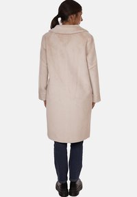 Cappotto in pelliccia sintetica beige con colletto classico, maniche lunghe e taglio dritto, che arriva a metà coscia. Indossato sopra jeans scuri e stivaletti neri.