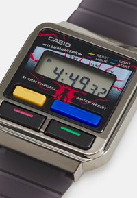 Casio digital klokke med sølvrektangulært hus, mørk gummirem, LCD-skjerm som viser "4:49.32," fargerike knapper i gul, blå, grønn og rosa.