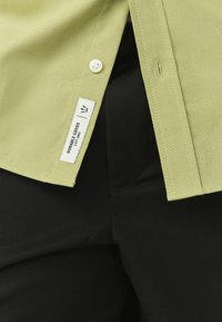 Oliiviroheline särk, millel on valge nööp ja silt tekstiga "Durable Goods Lot 082" mustade pükste kohal.