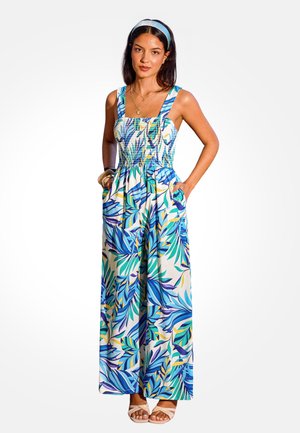 Kobieta w bezrękawniku i szerokich spodniach typu jumpsuit w tropikalny wzór liści w kolorach niebieskim, zielonym i żółtym, w sandałach, z opaską na głowie i warstwową biżuterią.