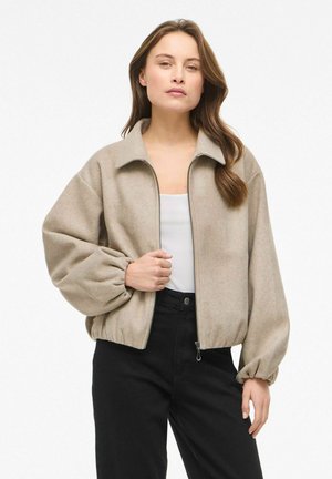 Naine, kes kannab beežikat lukuga jopet elastsete mansettidega, valget topi ja kõrge vöökohaga musti pükse, seisab ühevärvilise valge tausta ees.