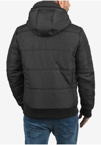 Schwarze Steppjacke mit Kapuze, aus strukturiertem Stoff, mit gerippten Bündchen und tailliertem Schnitt. Sichtbares Steppmuster auf der Rückseite.