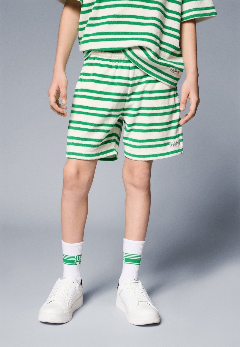 Enfant portant un short rayé vert et blanc assorti à un t-shirt, des chaussettes blanches avec des rayures vertes et des baskets blanches, sur un fond uni.