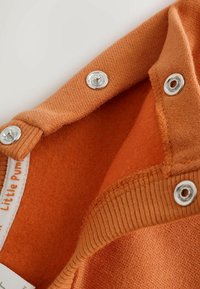 Close-up of orange fabric with silver snap buttons and ribbed edges, showing the interior soft texture and part of a white clothing label.