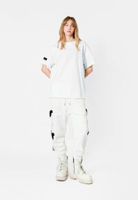 Camiseta blanca de gran tamaño con un sutil parche en el frente, combinada con pantalones cargo blancos que presentan aplicaciones florales negras y botas gruesas blancas.