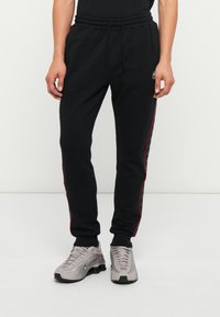 Pantalones jogger negros hechos de tela suave con rayas laterales rojas, que presentan una cinturilla elástica y un cordón, combinados con zapatillas gris.